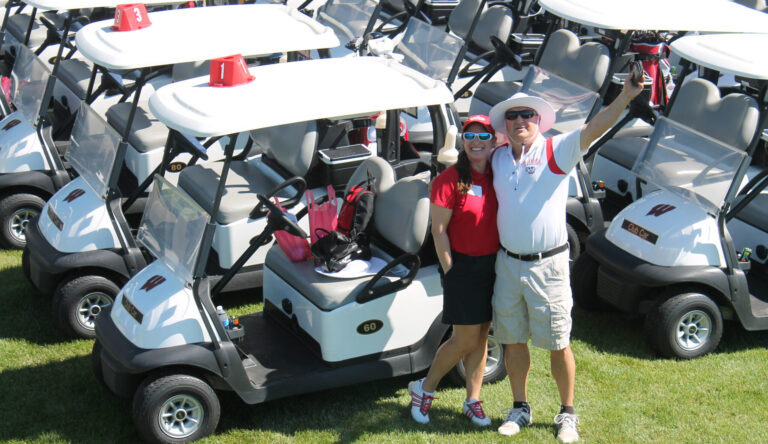 Alumni waving in front of a flock of golf carts