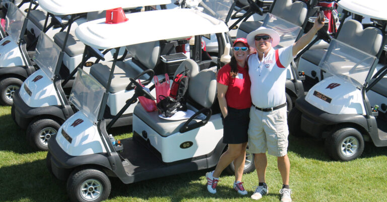 Alumni waving in front of a flock of golf carts