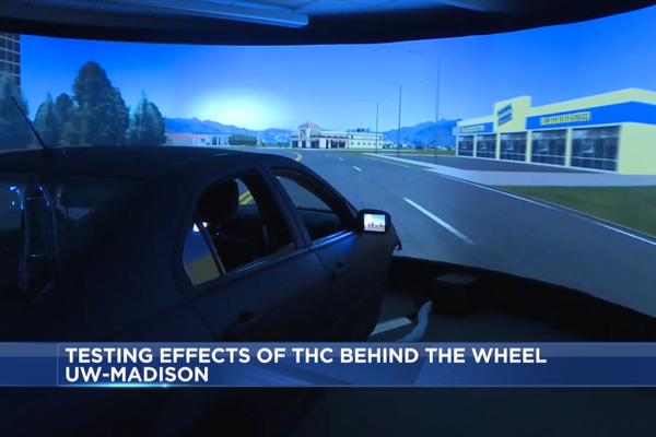 An image of a driving simulator from a newscast. In the foreground, the body of a car. In the background, a large screen with a simulated road.