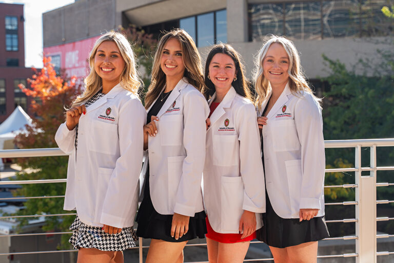 Four female PharmD students pose outside in their white coats, holding their lapels to showcase the School of Pharmacy logo.