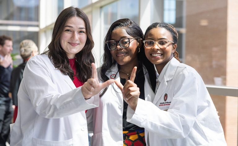 Three students in white coats pose with their hands in a W shape during the Pinning Ceremony